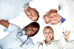 Picture of two Physicians (male and female) and two Nurses (male and female) all standing in a circle hugging while looking down at the ground smiling.