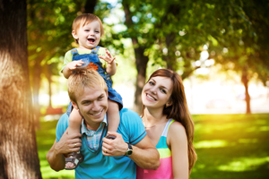 Picture of a young couple walking around in a park. There is a baby boy on the dad&apos;s shoulders and they are all happy and smiling.