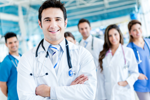 Picture of a Male Physician crossing his arms and smiling. He is wearing a medical coat with a stethoscope around his neck. There is a blurred background of two Physicians (male and female) and two Nurses (male and female) smiling as well.