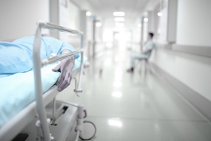 Picture of a hospital hallway with half of a swing bed in the picture and a person in the background sitting in a chair in the hallway (faded background)