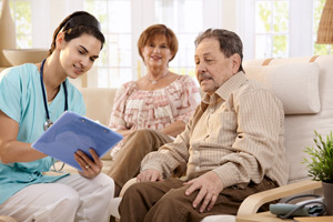 Picture of a Home Care Nurse sitting down talking to an elderly couple while holding a clipboard and pen in her hand