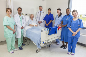 Picture of Medical Professionals (2 males and 5 females) standing in a room with a female patient that is sitting up in a swing bed