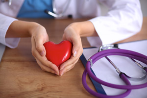 Picture of a female Physician's hands holding a heart object out and forming her hands into a heart. There is a heart shaped stethoscope on top of a clipboard on a desk next to her