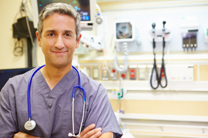 Picture of a male Nurse standing in a hospital room folding his arms and smiling. He has a stethoscope around his neck.